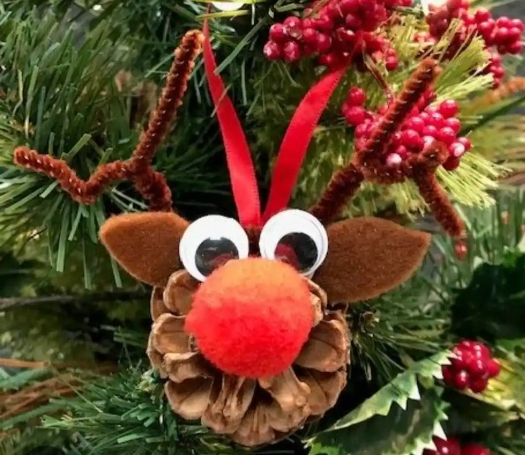 A handmade reindeer ornament made from a pinecone, brown felt ears and antlers, googly eyes, and a red pom-pom nose, hanging…
