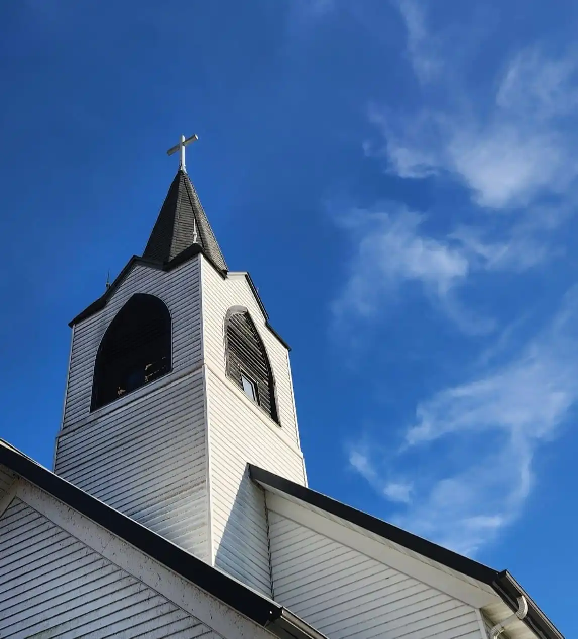 A low-angle photo shows the white wooden steeple of a church against a bright blue sky with wispy clouds. The steeple has a…