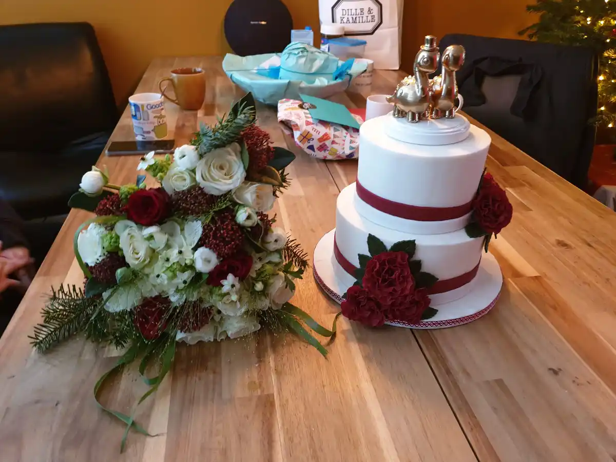 A two-tiered white wedding cake with red ribbon and red roses, topped with gold elephant figurines, sits on a wooden table…