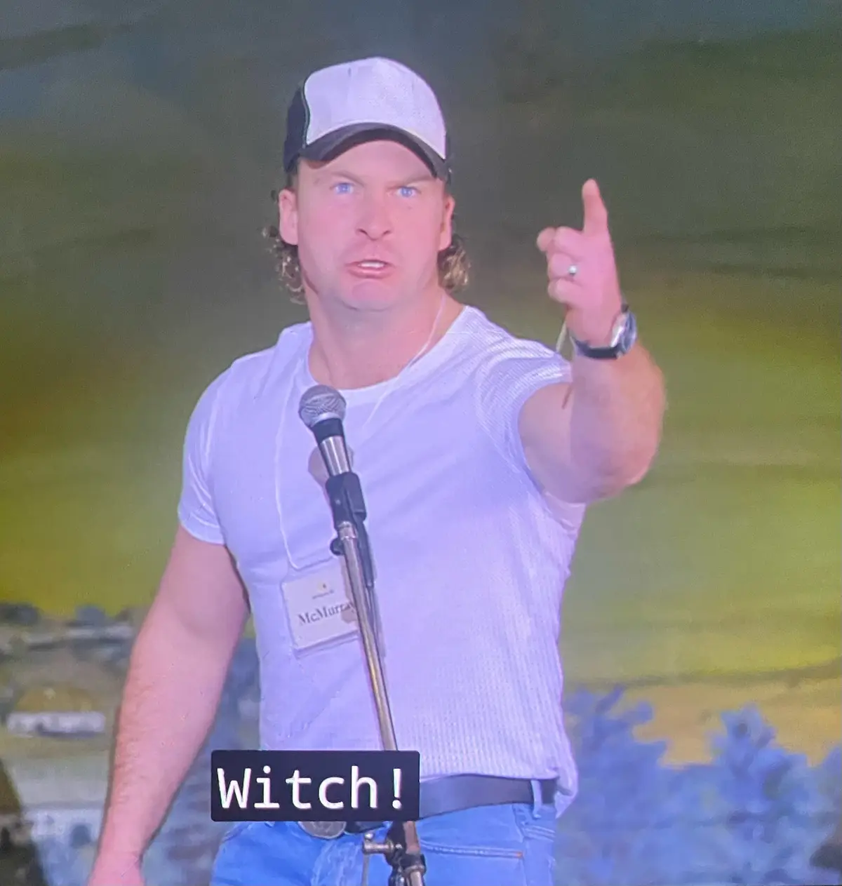 A man with blue eyes and curly hair, wearing a white t-shirt, blue jeans, and a white-and-black baseball cap, stands at a…