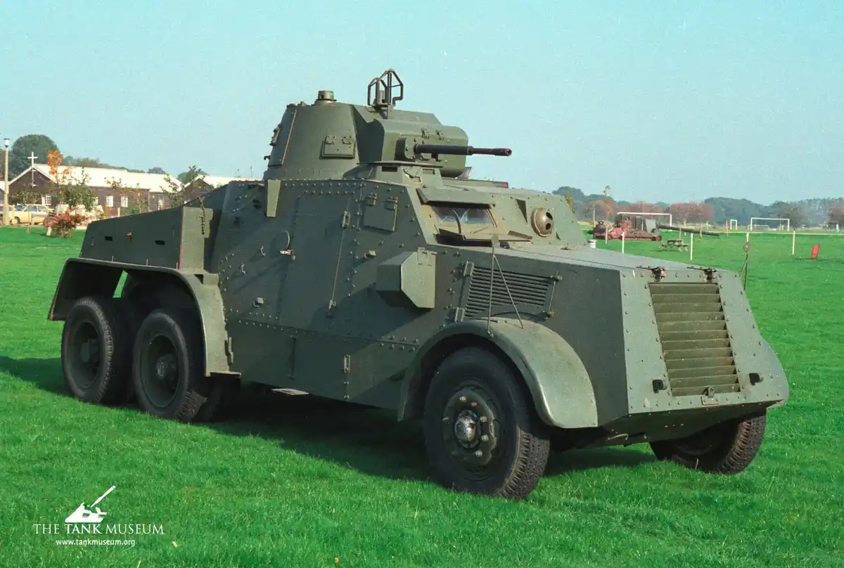 A green armored vehicle, resembling a World War II-era armored car, is parked on a grassy field with a museum building in…