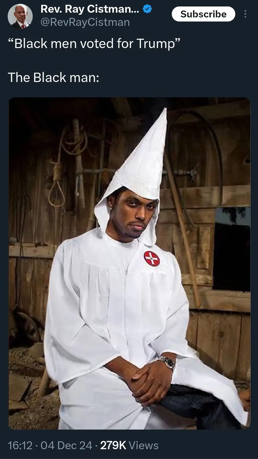 A Black man sits in a rustic wooden room wearing a white Ku Klux Klan hood and robe with a red cross emblem on the chest…