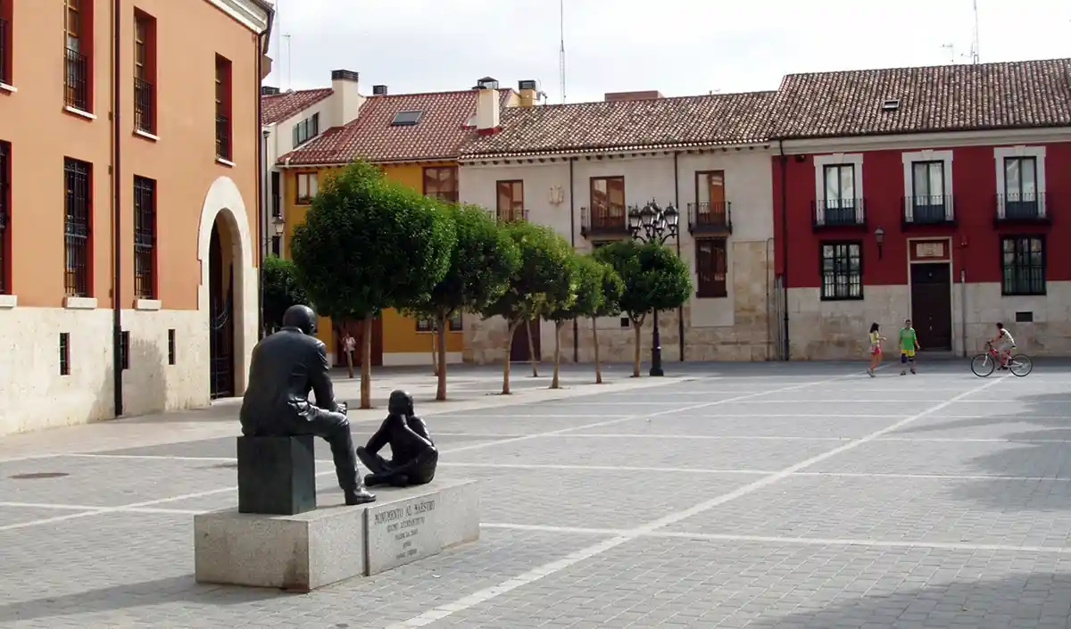 A public square features a bronze statue of a seated adult and a smaller child, both in contemplative poses, on a stone…