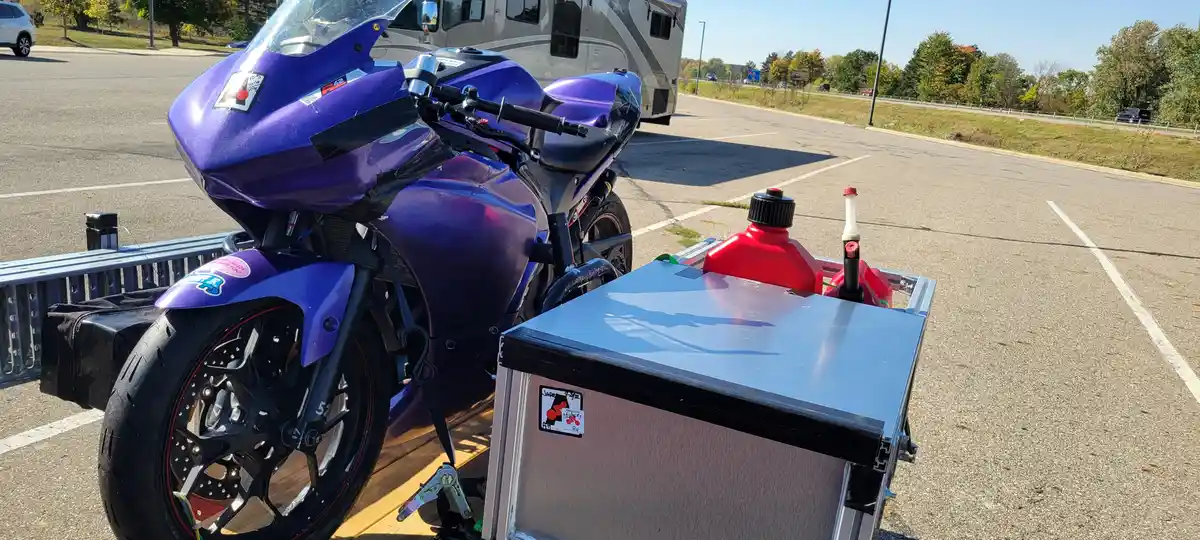 A shiny purple sport motorcycle is parked on a trailer in a parking lot, next to a blue toolbox with two red fuel containers…