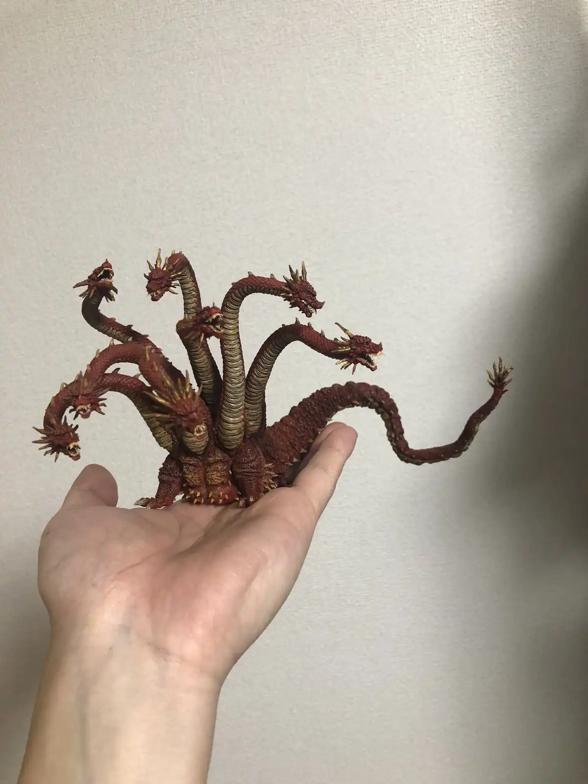 A person's hand holds a small, intricately detailed figurine of a multi-headed red dragon with golden horns and spines, set…