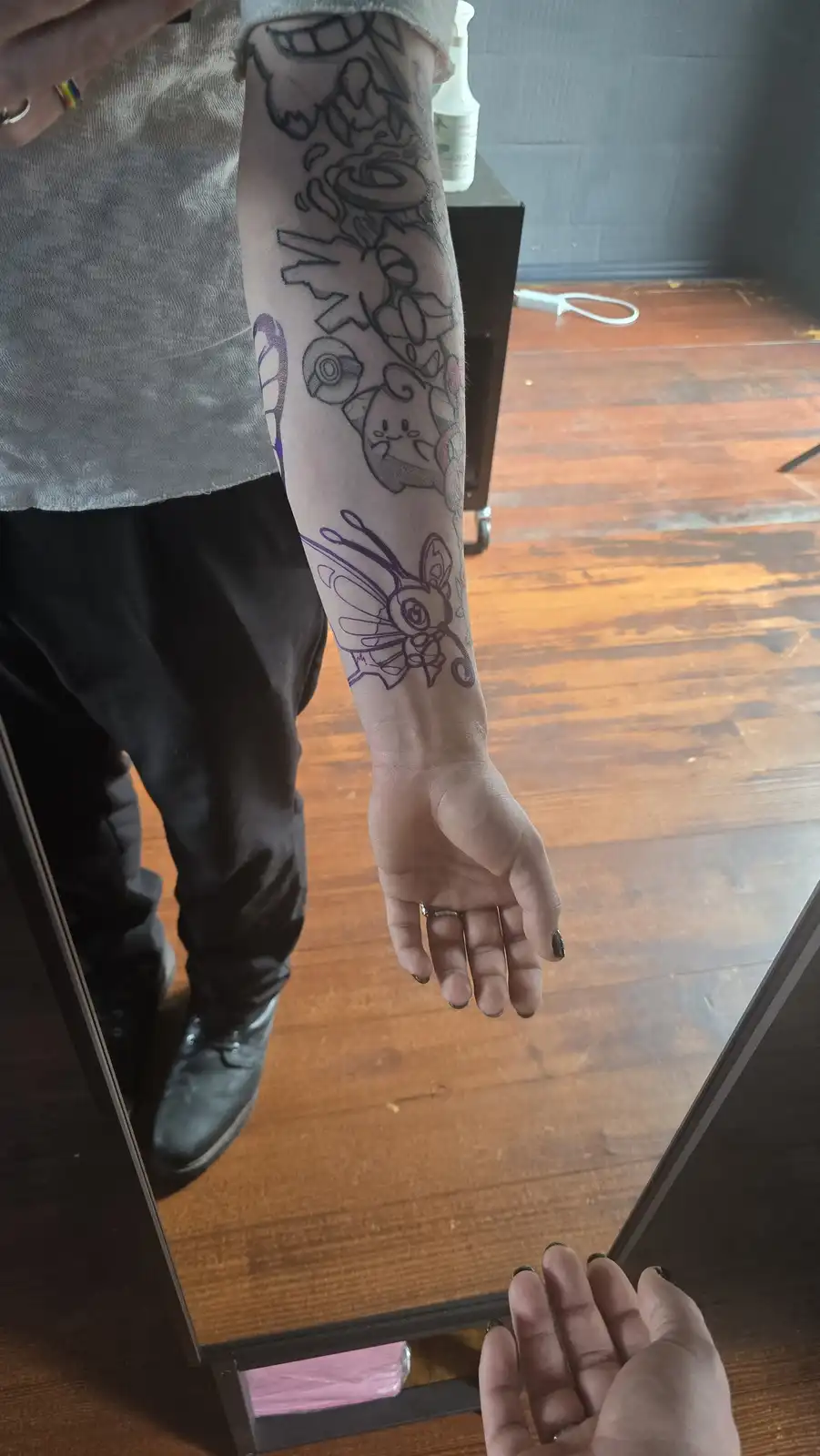 A person's left arm is shown in a mirror, displaying a partially completed tattoo sleeve with various Pokémon characters…