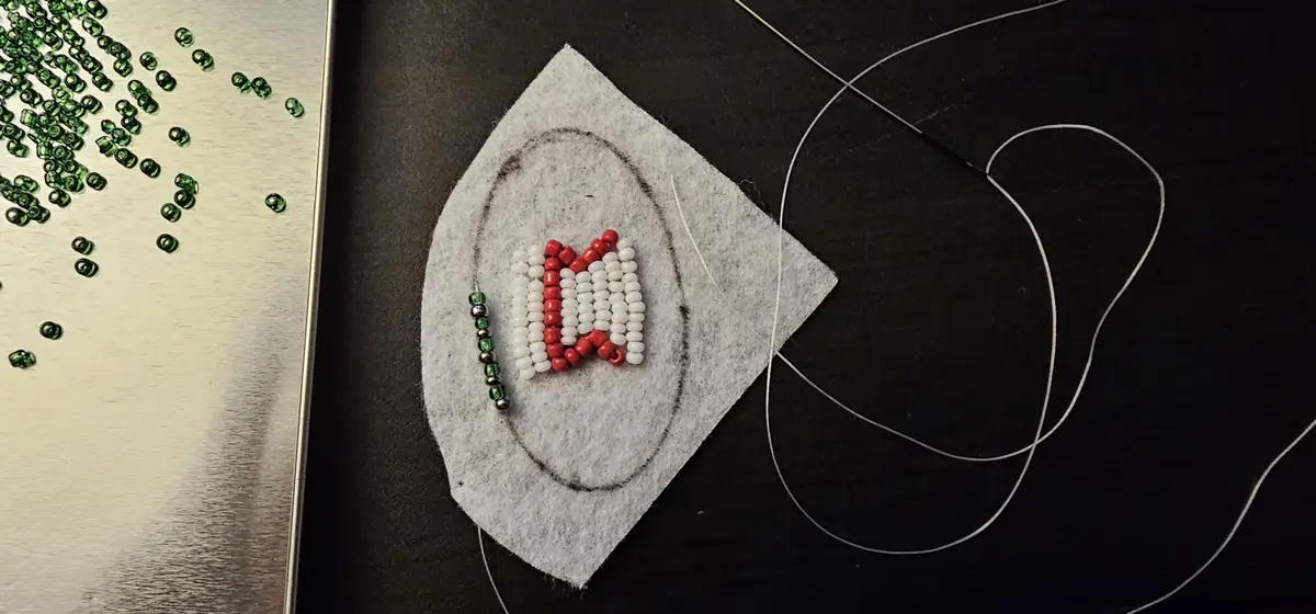 A close-up photo shows a beading project in progress on a gray felt square with a pencil outline. Red and white beads form a…