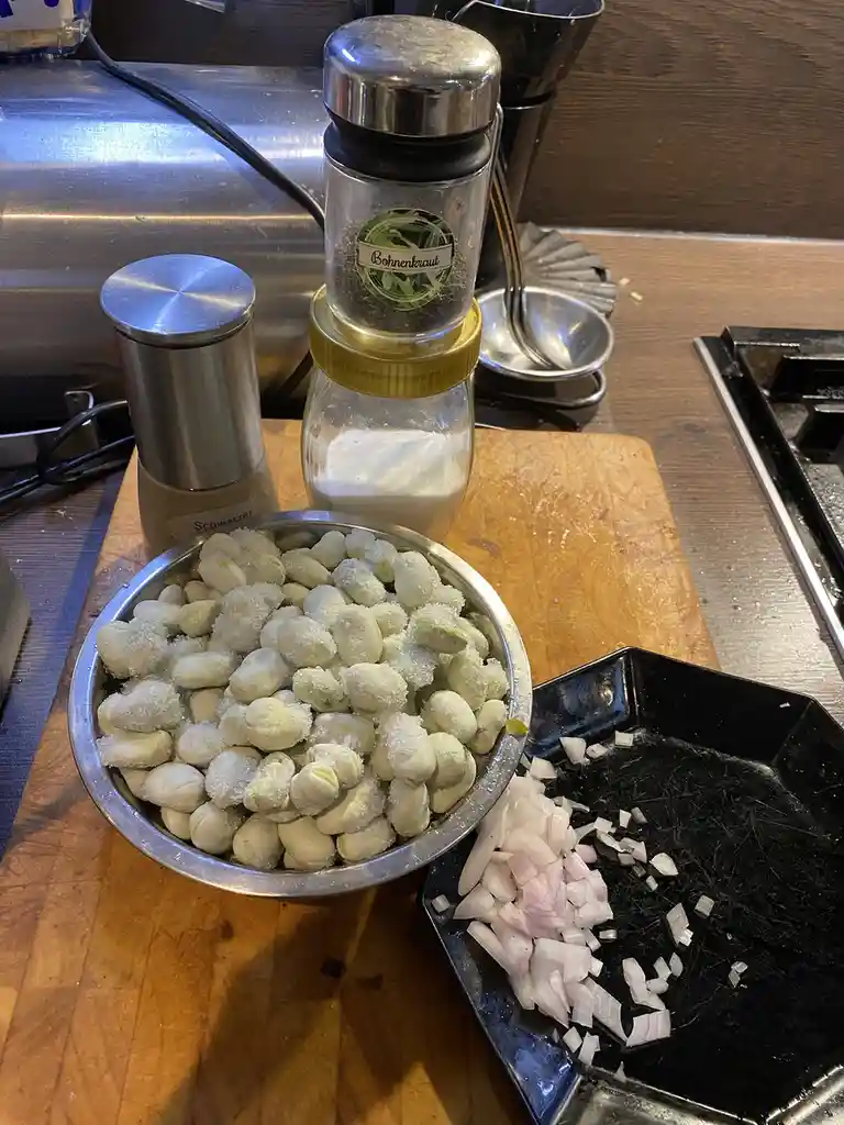 A kitchen counter shows a metal bowl of salted fava beans, a black tray with chopped onions, and stacked spice jars labeled…