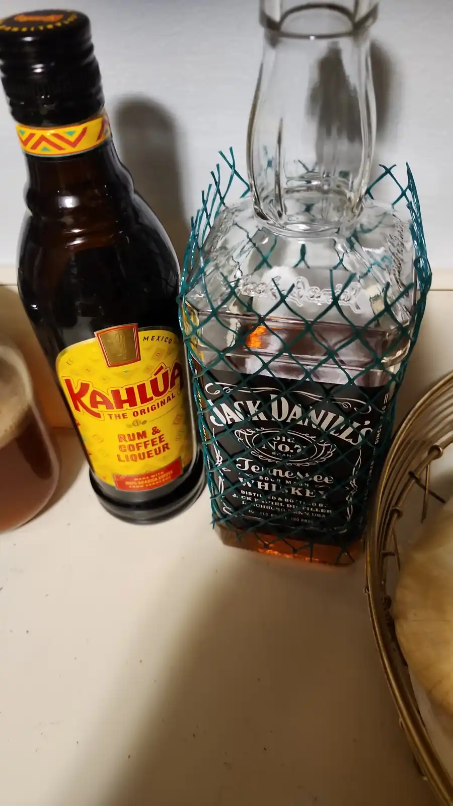 A bottle of Jack Daniel's Tennessee Whiskey is wrapped in a green wire mesh cage, standing next to a bottle of Kahlúa rum…