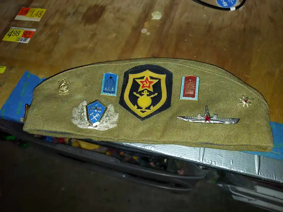 A khaki-colored military cap is displayed on a wooden surface, covered with numerous Soviet-era military insignia including…