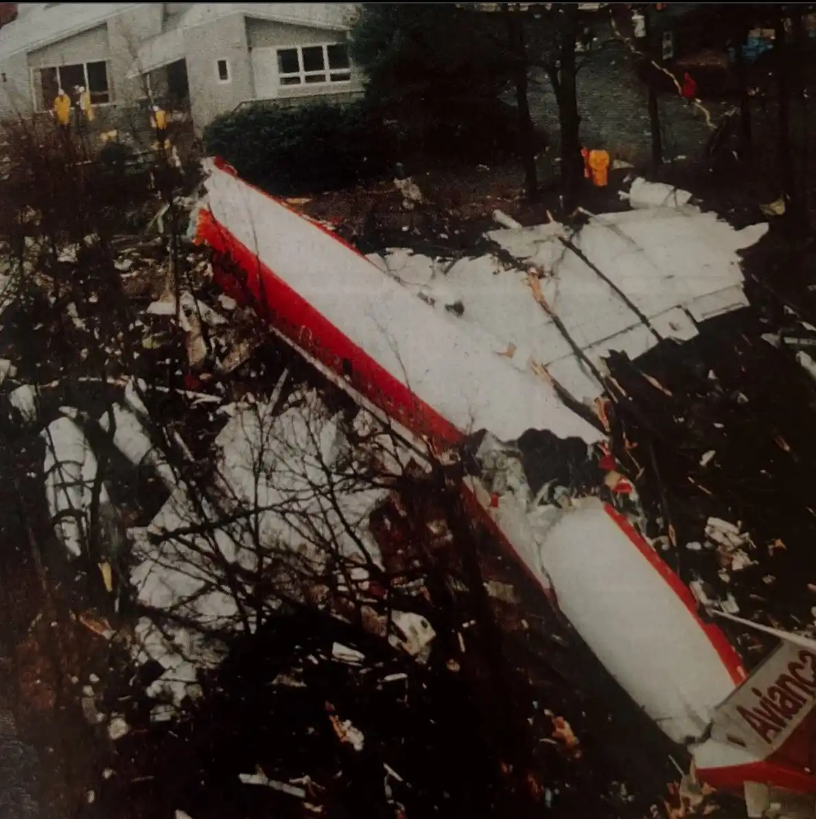 A severely damaged airplane with red and white livery lies in pieces across a wooded area near buildings, with emergency…