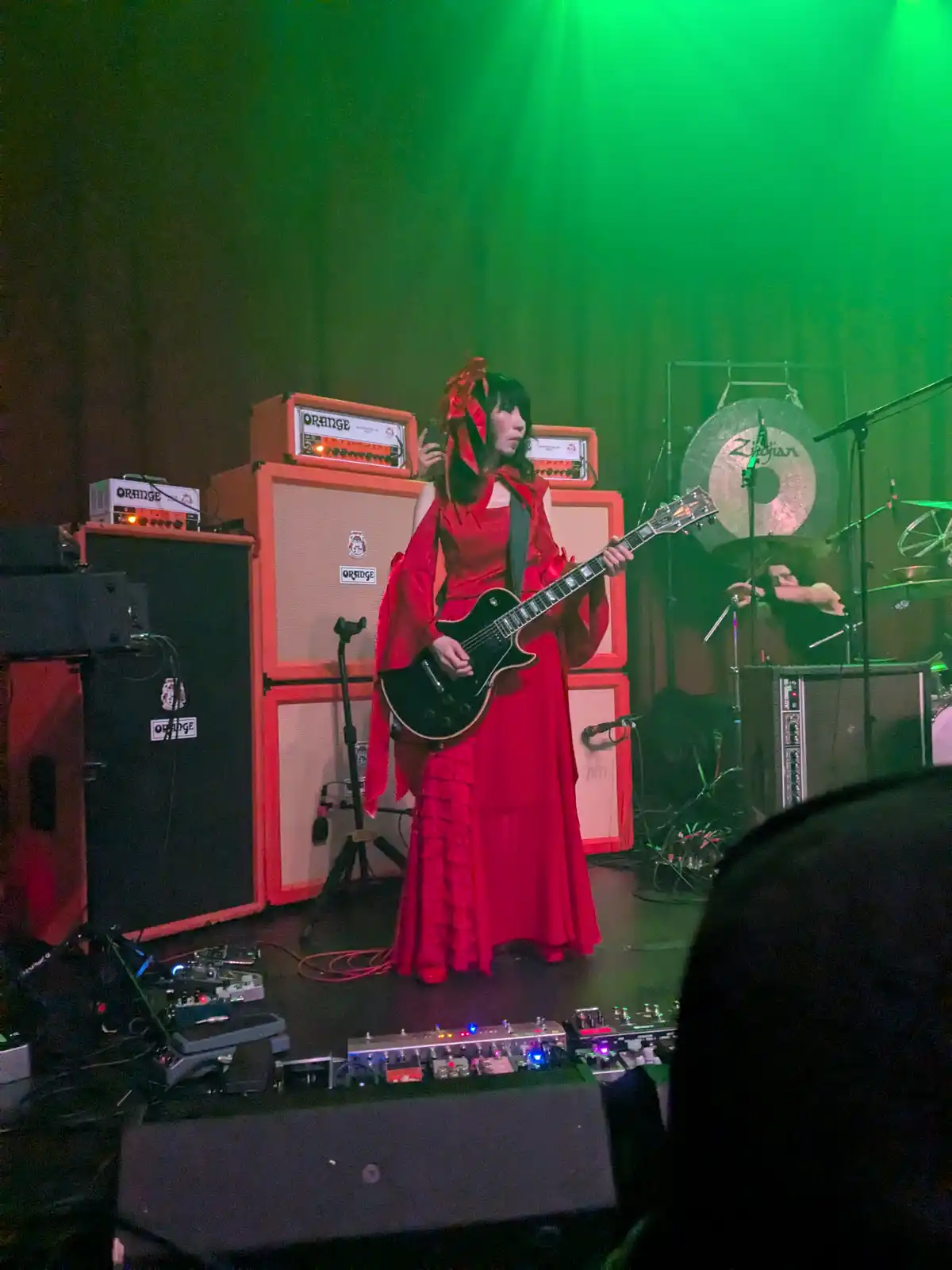 A woman in a red dress and red hair accessories plays a black electric guitar on a stage bathed in green light, surrounded…