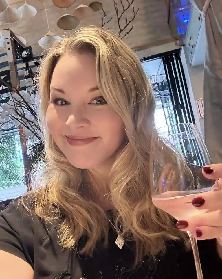 A woman with blonde hair and red nail polish holds a wine glass while smiling at the camera in what appears to be a…