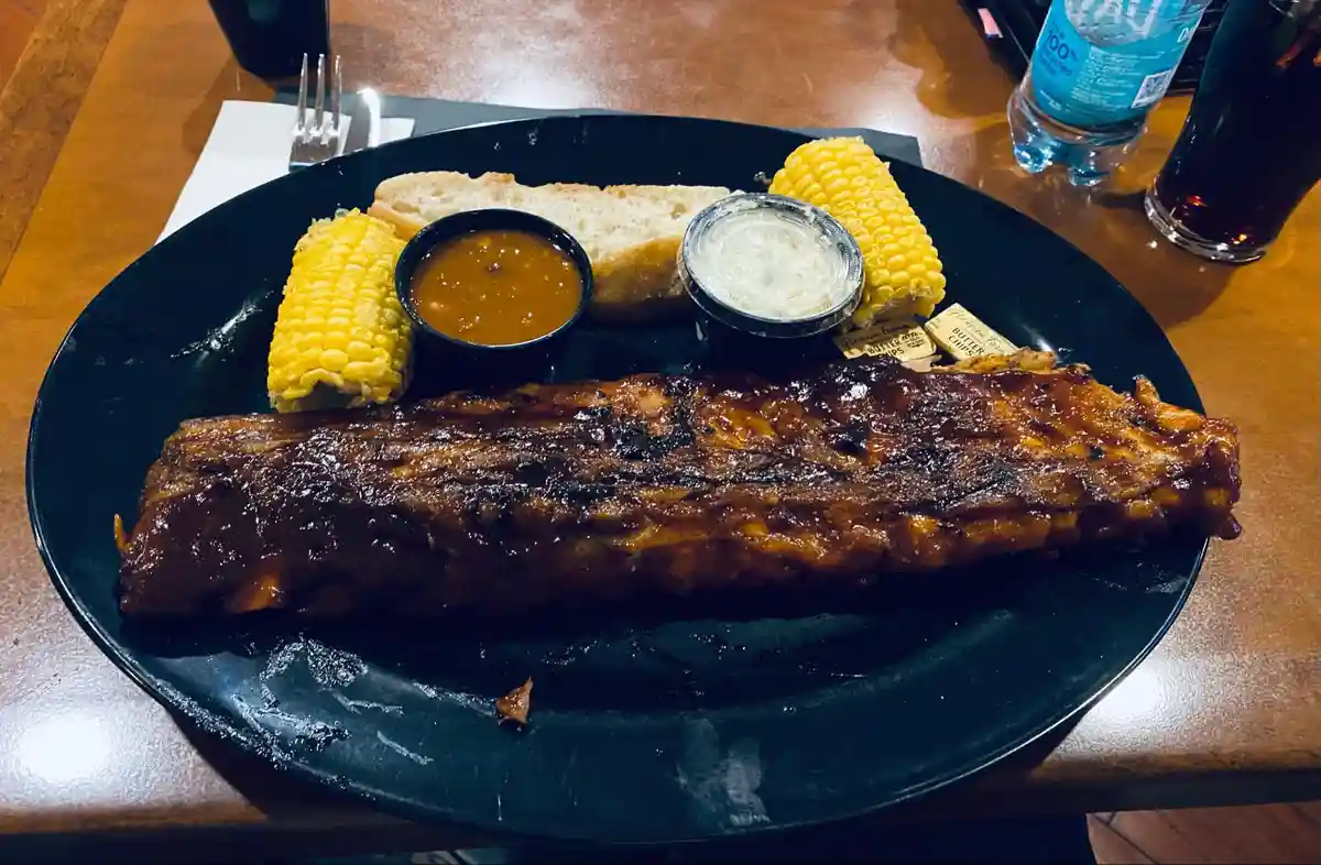 A close-up photo of a large, glossy rack of barbecue ribs on a black plate, accompanied by two ears of corn on the cob, a…