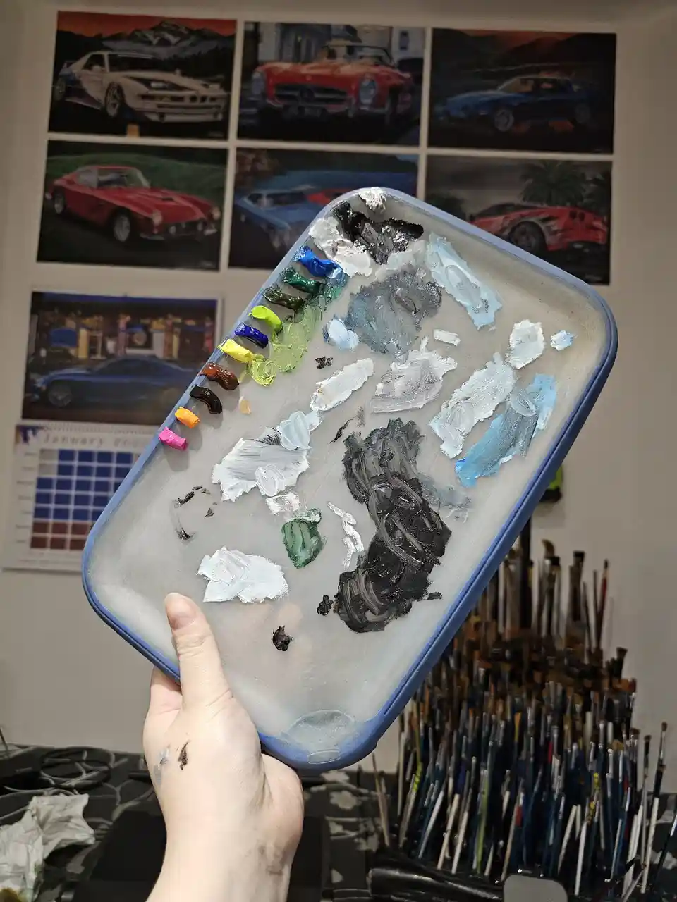 A hand holds up a heavily used paint palette with smears of various colors, including large patches of black and white…