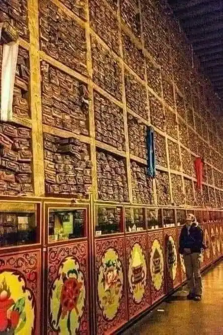 A person stands in awe before a massive, towering wall covered in countless small, ornate, wooden prayer wheels or religious…