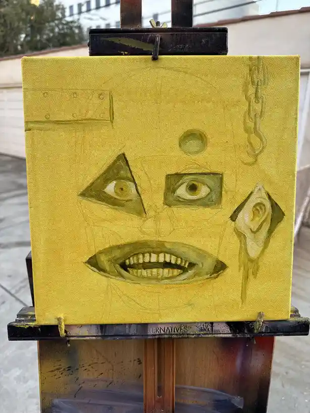 A yellow canvas painting on an easel depicts a surreal, distorted face with one triangular eye, one rectangular eye, a wide…