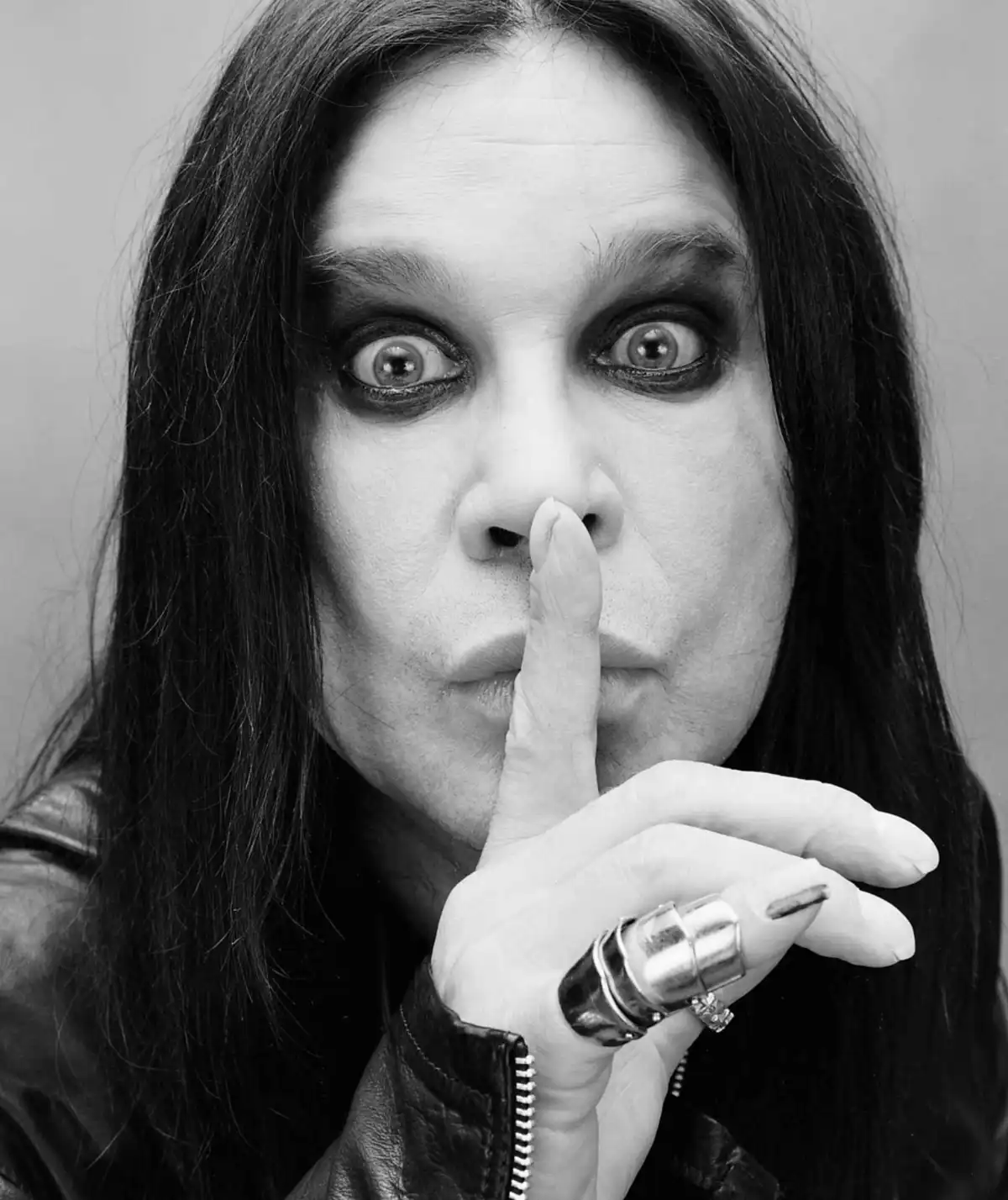 A black-and-white photo of Ozzy Osbourne making a 'shhh' gesture with his finger over his lips, his eyes wide open, wearing…