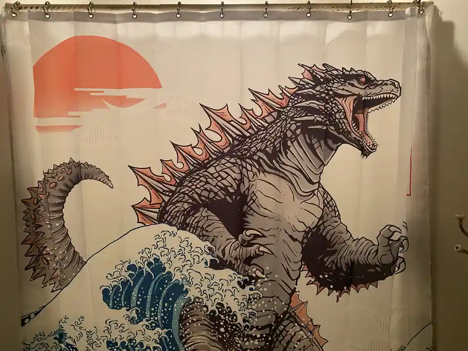 A shower curtain features a stylized Godzilla standing atop a large blue wave, with an orange sun in the background. The…