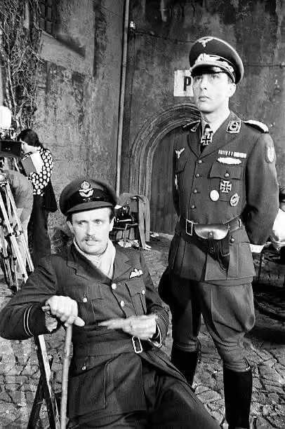 A black-and-white photo shows a man in a military uniform seated and holding a cane, while another man in a Nazi uniform…