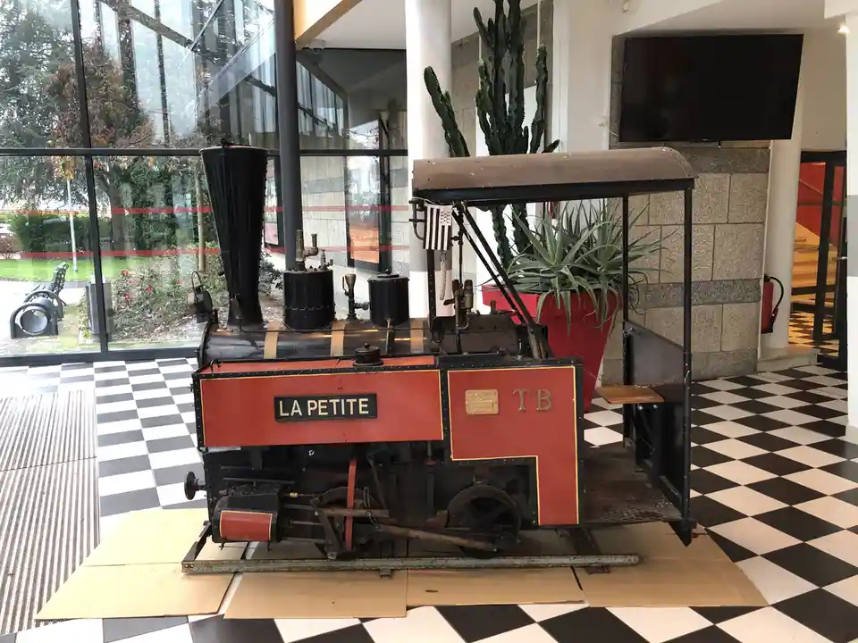 A small, red and black vintage steam locomotive named 'LA PETITE' sits indoors on a black-and-white checkered floor…