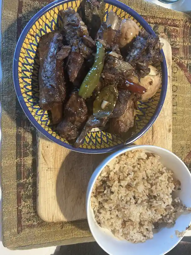 A top-down photo shows a bowl of dark, saucy meat with bones, potatoes, peppers, and bay leaves, next to a bowl of brown…