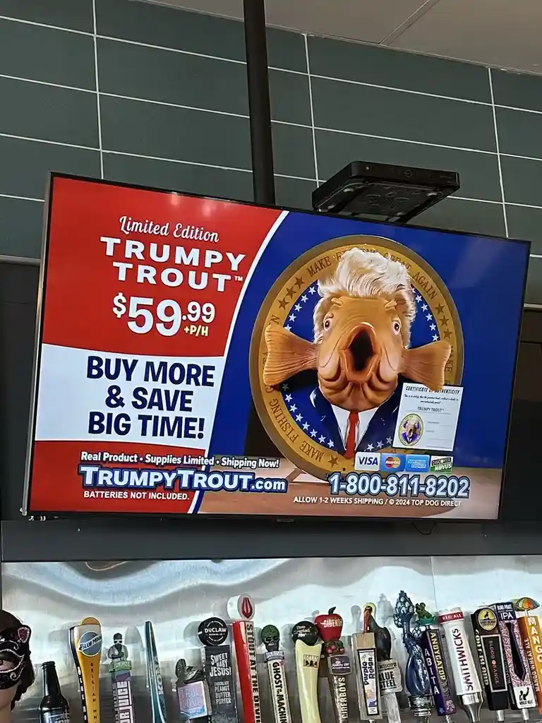 A TV screen in a bar displays a satirical advertisement for a 'Limited Edition TRUMPY TROUT™' priced at $59.99 plus…
