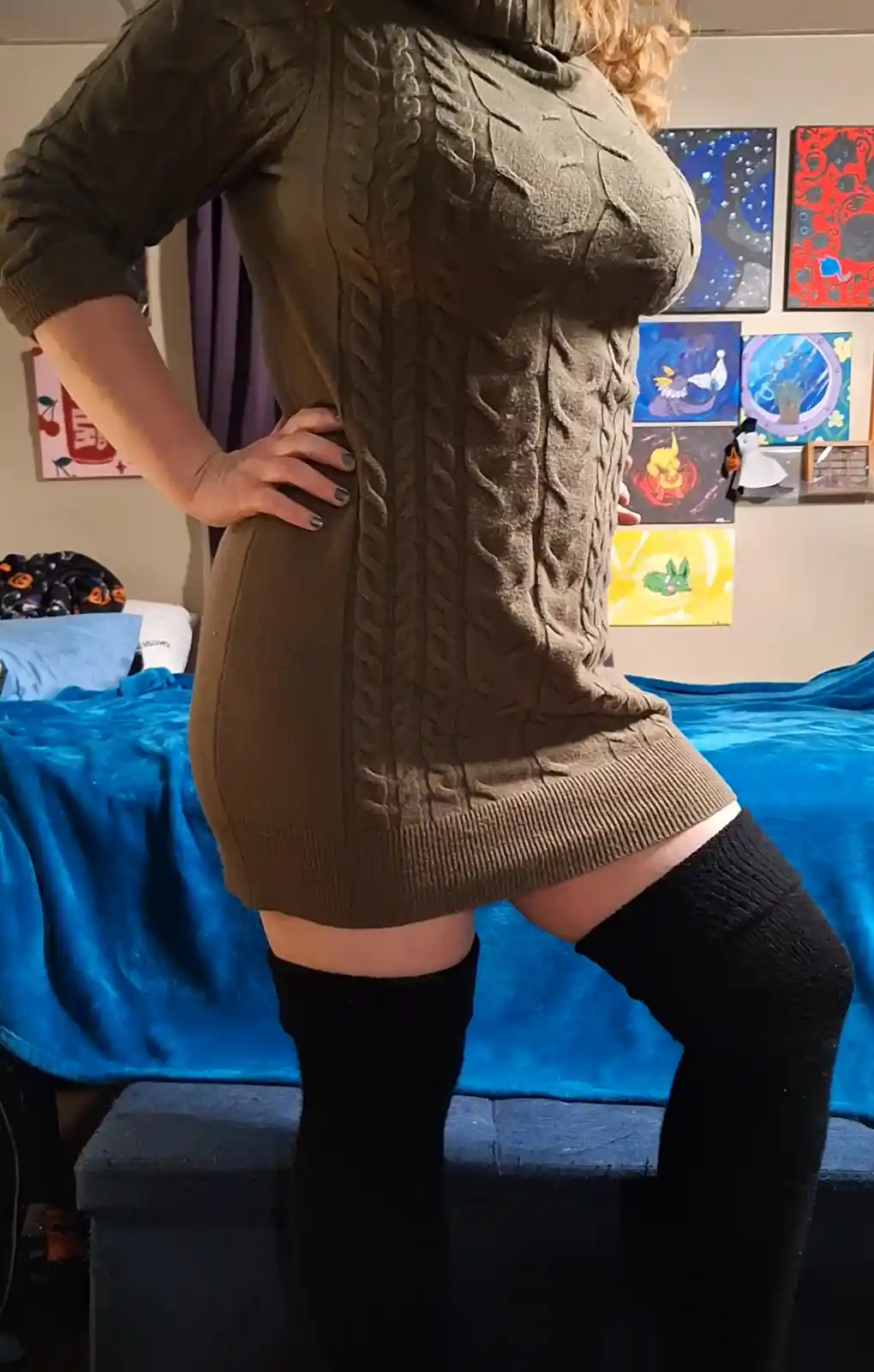 A person stands in a bedroom, wearing a tight-fitting olive green cable-knit sweater dress that accentuates their curves…