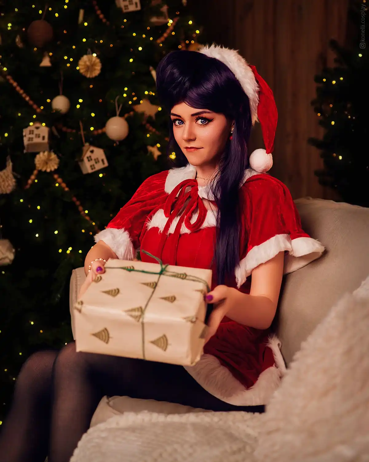 A woman with dark hair and blue eyes, wearing a red and white Santa outfit with a matching hat, sits on a couch holding a…