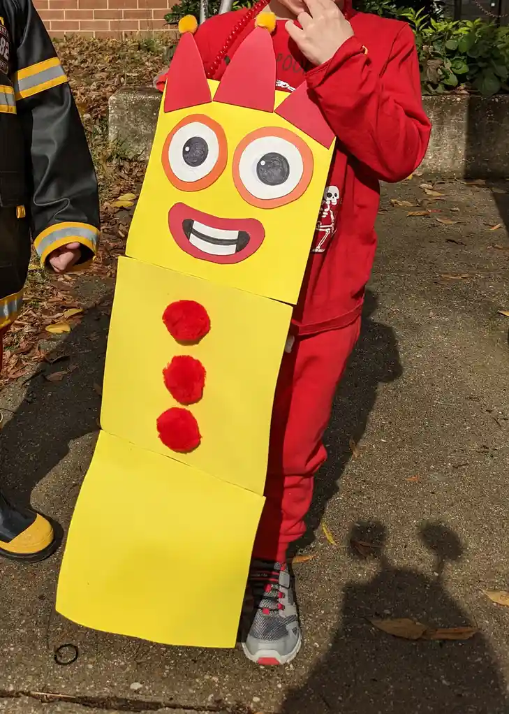 A child in a red outfit is wearing a homemade yellow cardboard costume resembling a robot with large cartoon eyes, a smiling…
