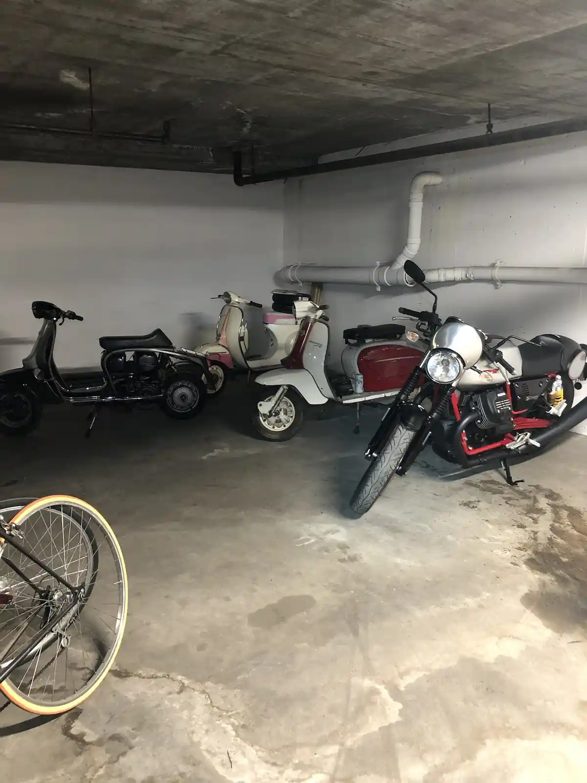 A dimly lit concrete garage contains several scooters and one motorcycle parked in a row against the wall, with a bicycle…