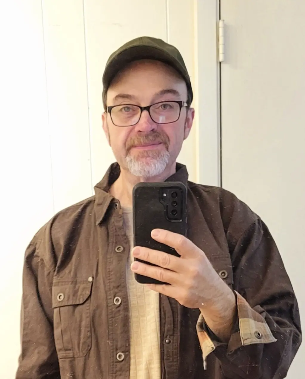A middle-aged man with glasses and a beard takes a mirror selfie holding a black smartphone. He wears a dark brown button-up…