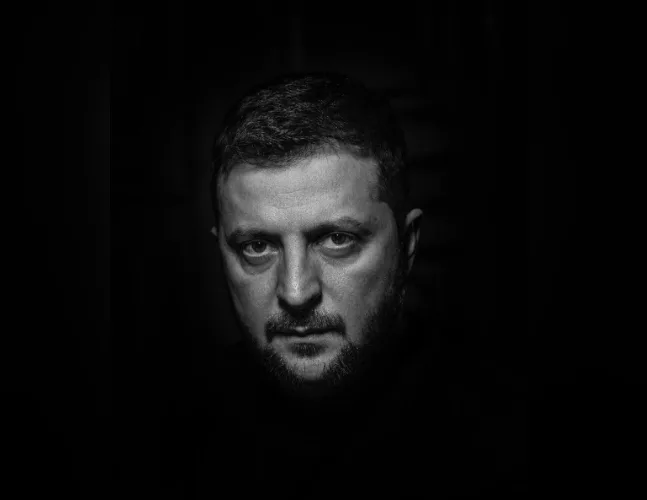 A black-and-white portrait of a man with short dark hair and a beard, looking intensely at the camera with a serious…
