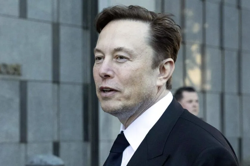 A man in a dark suit and tie, identified as Elon Musk, is shown in profile outdoors against a modern building facade. He…