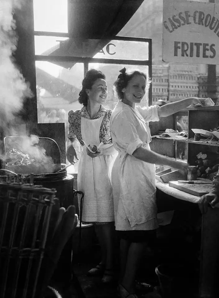 A vintage black-and-white photo shows two smiling women in aprons working at a French street food stall, with steam rising…