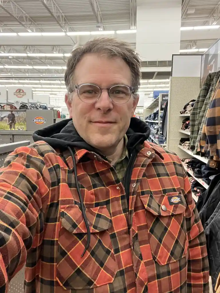 A man with glasses and a slight smile takes a selfie in a retail store, wearing a red, black, and brown plaid Dickies shirt…