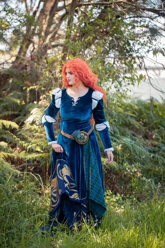 A person with bright red curly hair stands in a grassy, wooded area wearing a blue velvet dress with white puffy sleeves, a…