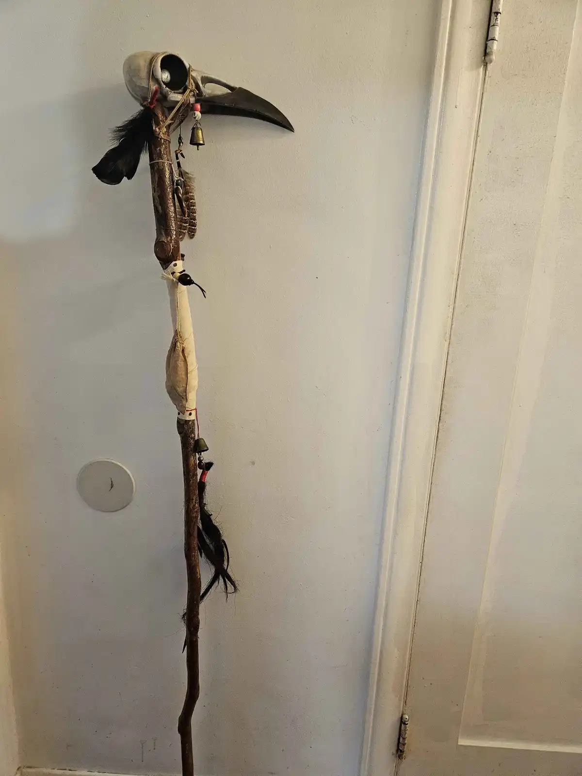 A rustic walking stick with a bird skull at the top, adorned with feathers, bells, and a cloth-wrapped section, leans…