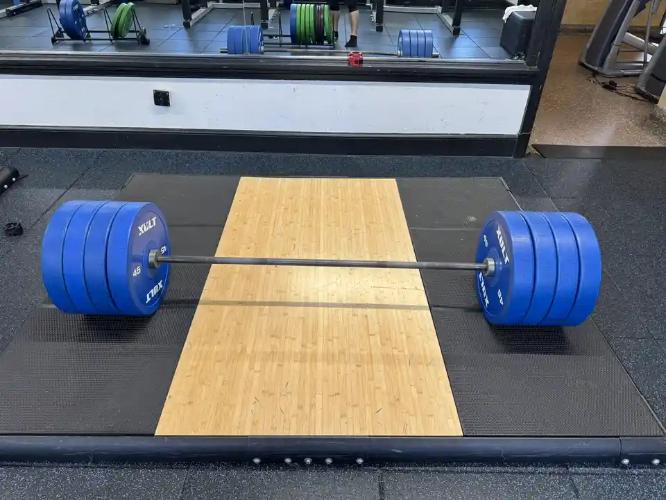 A heavy barbell with blue 45lb plates rests on a wooden lifting platform in a gym, with no person visible nearby. The…