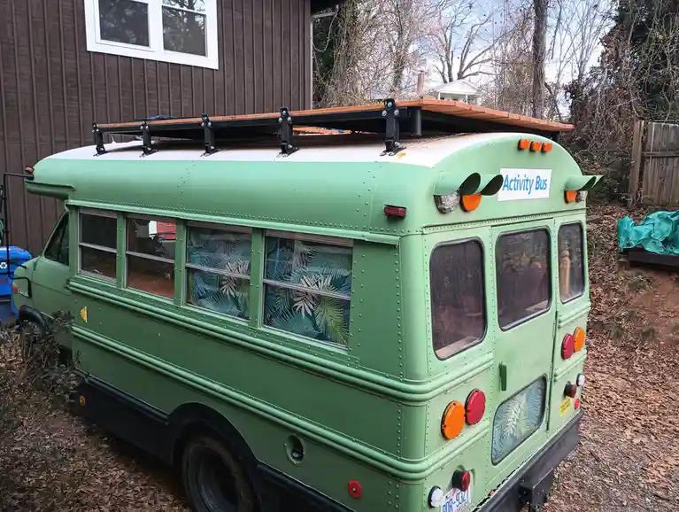 A green, vintage-style school bus parked in a yard, modified with a wooden rooftop deck and tropical palm leaf curtains…