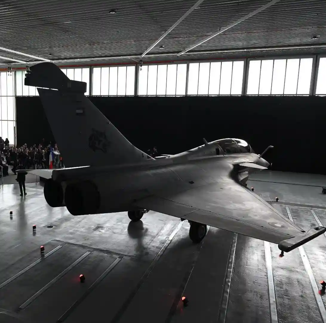 A grey military jet, likely a Dassault Rafale, is displayed indoors on a polished floor with red spotlights, facing away…