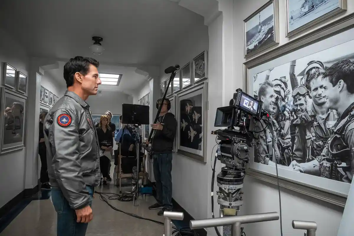 A man in a gray flight jacket with a U.S. Navy patch stands in a hallway lined with framed black-and-white photos of pilots…