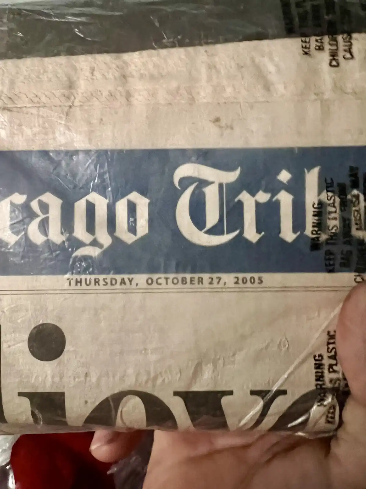 A close-up photo shows a hand holding a newspaper wrapped in plastic, with the Chicago Tribune masthead visible and the date…