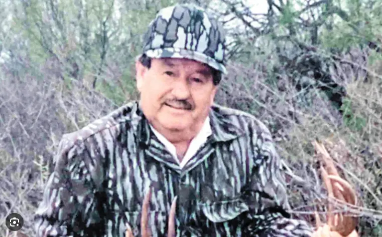 An older man with a mustache wearing a camouflage hat and jacket stands outdoors in a wooded area, holding a set of deer…