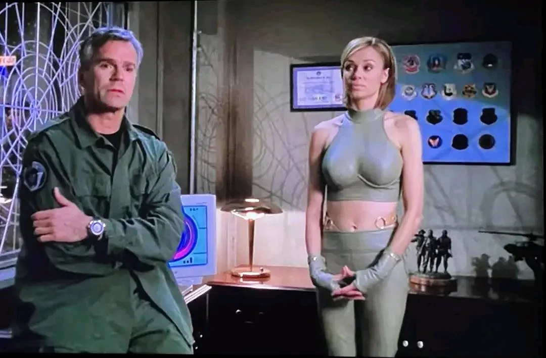 A man in a green military-style uniform stands with arms crossed, looking skeptical, while a woman in a tight, form-fitting…