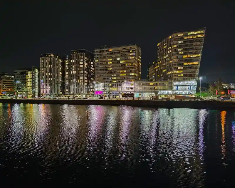 A nighttime cityscape of modern buildings along a waterfront, their lights reflecting in rippling water. The overlaid text…