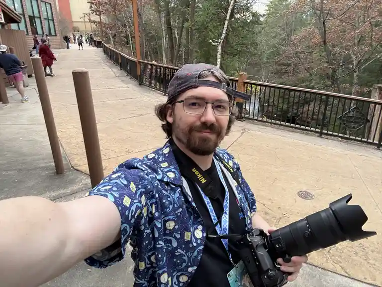 A man with glasses and a backwards cap takes a selfie holding a large DSLR camera with a telephoto lens. He wears a colorful…