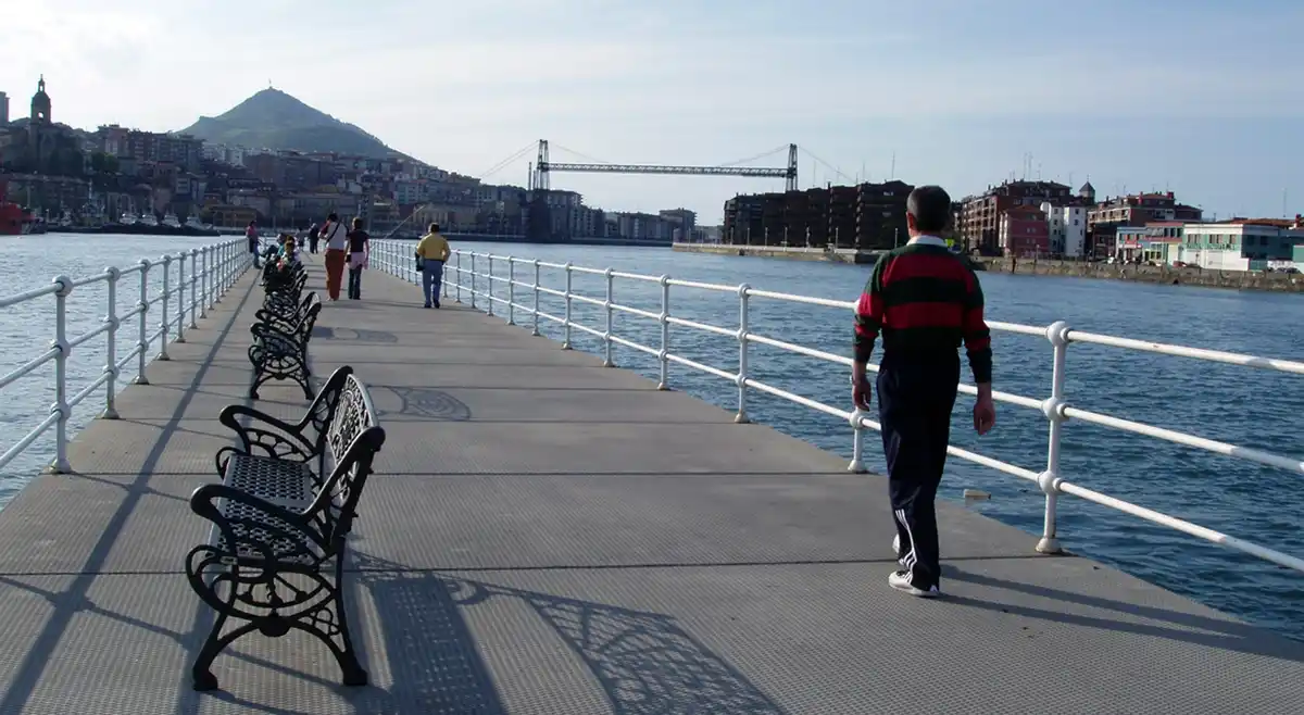 A man in a red and green striped sweater walks away down a long pier lined with empty ornate benches, overlooking water with…