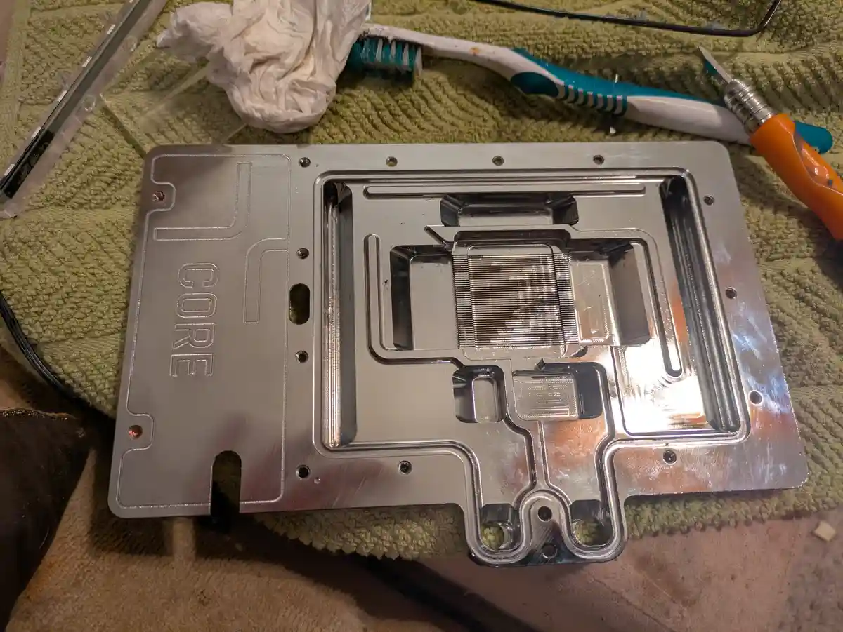 A close-up photo shows a cleaned, shiny silver CPU water block with 'CORE' engraved on its side, resting on a green towel…