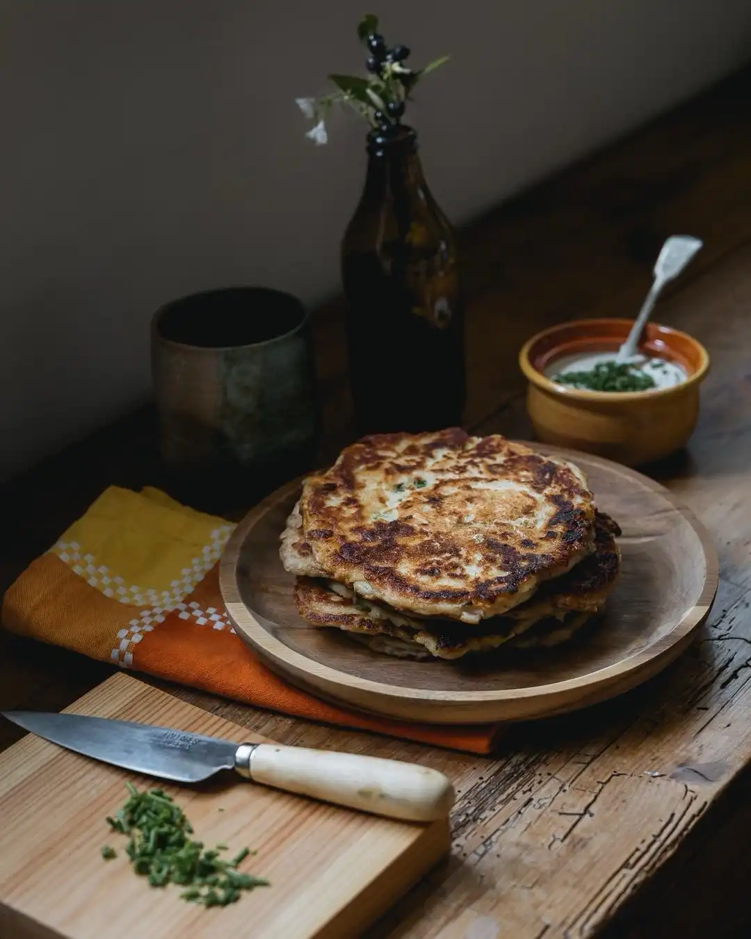 A rustic food photograph shows a stack of golden-brown, slightly charred pancakes on a wooden plate, set on a wooden table…